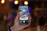 A hand holding a smartphone displays a photo of a modern cafe or restaurant setting. The screen shows people seated at tables, with a vibrant and lively atmosphere suggesting a social gathering or casual meeting. The background is blurred with warm, ambient lighting.