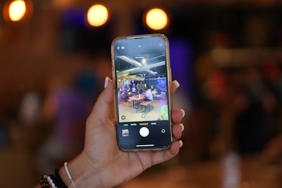 A hand holding a smartphone displays a photo of a modern cafe or restaurant setting. The screen shows people seated at tables, with a vibrant and lively atmosphere suggesting a social gathering or casual meeting. The background is blurred with warm, ambient lighting.