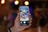 A hand holding a smartphone displays a photo of a modern cafe or restaurant setting. The screen shows people seated at tables, with a vibrant and lively atmosphere suggesting a social gathering or casual meeting. The background is blurred with warm, ambient lighting.