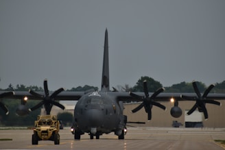 A large military aircraft with multiple propellers is situated on an airfield runway, facing forward. A smaller military vehicle is positioned in front of the aircraft. The sky appears overcast, and trees can be seen in the background along with some hangars.