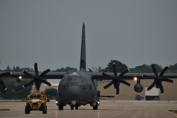 A large military aircraft with multiple propellers is situated on an airfield runway, facing forward. A smaller military vehicle is positioned in front of the aircraft. The sky appears overcast, and trees can be seen in the background along with some hangars.