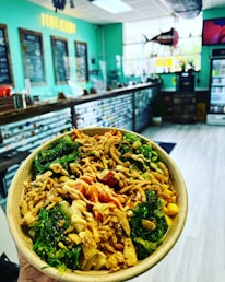 A vibrant food bowl with a mix of fresh vegetables, crispy toppings, and sauce. The background features a modern, relaxed cafe setting with a turquoise color scheme, menu boards, and a sign reading 'I LIKE IT RAW'.