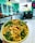 A vibrant food bowl with a mix of fresh vegetables, crispy toppings, and sauce. The background features a modern, relaxed cafe setting with a turquoise color scheme, menu boards, and a sign reading 'I LIKE IT RAW'.
