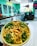 A vibrant food bowl with a mix of fresh vegetables, crispy toppings, and sauce. The background features a modern, relaxed cafe setting with a turquoise color scheme, menu boards, and a sign reading 'I LIKE IT RAW'.