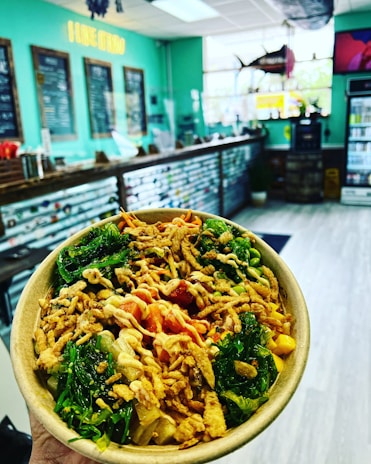 A vibrant food bowl with a mix of fresh vegetables, crispy toppings, and sauce. The background features a modern, relaxed cafe setting with a turquoise color scheme, menu boards, and a sign reading 'I LIKE IT RAW'.