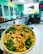 A vibrant food bowl with a mix of fresh vegetables, crispy toppings, and sauce. The background features a modern, relaxed cafe setting with a turquoise color scheme, menu boards, and a sign reading 'I LIKE IT RAW'.