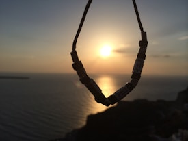 A beaded necklace is silhouetted against a sunset over the ocean. The sun casts a warm glow on the water, creating a serene and peaceful atmosphere. The horizon line is barely visible as the sky transitions from a deep blue to gold and orange hues.