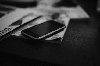A smartphone rests on top of a stack of magazines on a table. The setting appears to be dimly lit, creating a moody atmosphere.