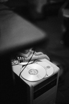 A stack of compact discs with handwritten labels is placed atop a desktop computer tower. A telephone handset with a coiled cord sits alongside the discs, adding a vintage feel. The image is in black and white, providing a nostalgic effect.