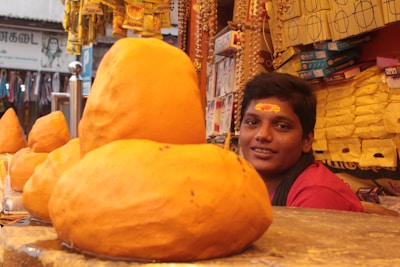 A young person with a tilak on their forehead is positioned behind a counter displaying large lumps of resting yellow-orange material, possibly turmeric. The background is filled with hanging strings of beads and various packaged goods, indicating a market or shop setting.