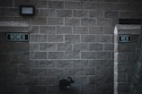 Outdoor restrooms with signs for women and men mounted on a masonry wall. The lighting is dim, creating a slightly somber atmosphere. A water fountain is visible beneath the men’s sign.