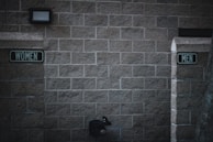 Outdoor restrooms with signs for women and men mounted on a masonry wall. The lighting is dim, creating a slightly somber atmosphere. A water fountain is visible beneath the men’s sign.