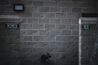 Outdoor restrooms with signs for women and men mounted on a masonry wall. The lighting is dim, creating a slightly somber atmosphere. A water fountain is visible beneath the men’s sign.