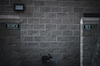 Outdoor restrooms with signs for women and men mounted on a masonry wall. The lighting is dim, creating a slightly somber atmosphere. A water fountain is visible beneath the men’s sign.