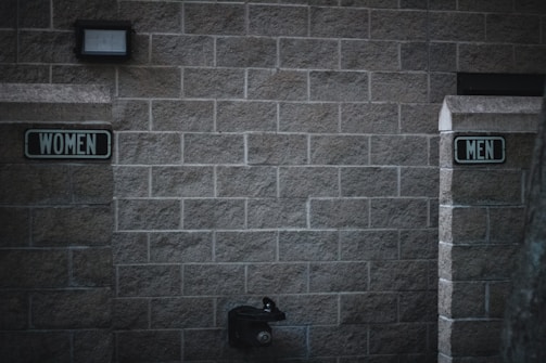 Outdoor restrooms with signs for women and men mounted on a masonry wall. The lighting is dim, creating a slightly somber atmosphere. A water fountain is visible beneath the men’s sign.