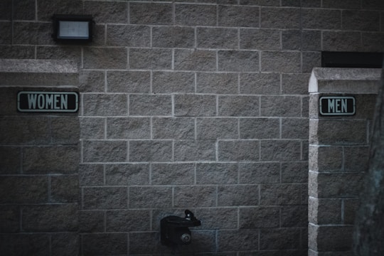 Outdoor restrooms with signs for women and men mounted on a masonry wall. The lighting is dim, creating a slightly somber atmosphere. A water fountain is visible beneath the men’s sign.