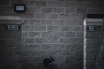 Outdoor restrooms with signs for women and men mounted on a masonry wall. The lighting is dim, creating a slightly somber atmosphere. A water fountain is visible beneath the men’s sign.