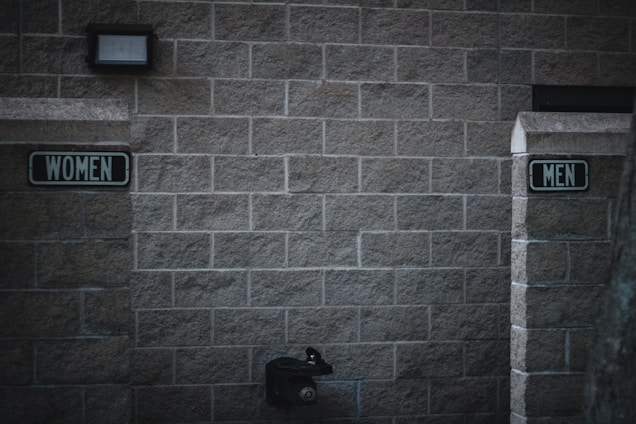 Outdoor restrooms with signs for women and men mounted on a masonry wall. The lighting is dim, creating a slightly somber atmosphere. A water fountain is visible beneath the men’s sign.