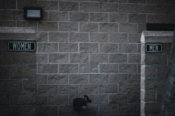 Outdoor restrooms with signs for women and men mounted on a masonry wall. The lighting is dim, creating a slightly somber atmosphere. A water fountain is visible beneath the men’s sign.