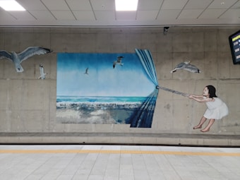 A mural on a concrete wall depicts a young child in a white dress pulling back a blue curtain to reveal an ocean scene. Several seagulls are flying across both the exposed ocean view and the wall outside of the mural, blurring the line between the painted and real world.