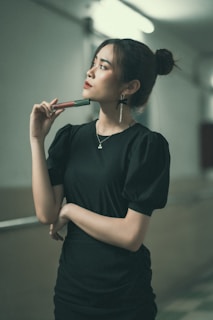 A woman stands holding a cosmetic product, gazing thoughtfully to the side. She wears a black dress with puffed sleeves and her hair is styled in a bun. The corridor setting is softly lit, creating a contemplative atmosphere.