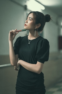 A woman stands holding a cosmetic product, gazing thoughtfully to the side. She wears a black dress with puffed sleeves and her hair is styled in a bun. The corridor setting is softly lit, creating a contemplative atmosphere.