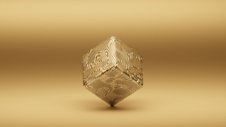 A geometric object floats against a uniform gold background. The object is a three-dimensional shape resembling a cube, but with intricate swirling patterns on its surface, giving it a textured appearance. The overall tone of the image is monochromatic with a minimalist style.