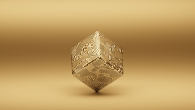 A geometric object floats against a uniform gold background. The object is a three-dimensional shape resembling a cube, but with intricate swirling patterns on its surface, giving it a textured appearance. The overall tone of the image is monochromatic with a minimalist style.