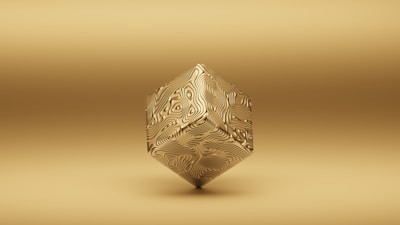 A geometric object floats against a uniform gold background. The object is a three-dimensional shape resembling a cube, but with intricate swirling patterns on its surface, giving it a textured appearance. The overall tone of the image is monochromatic with a minimalist style.