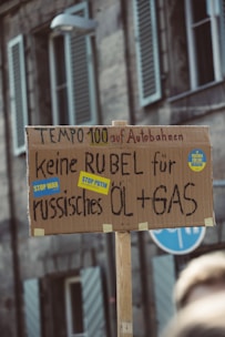 A protest sign made from cardboard with hand-written text in German. The background shows a blurry urban setting with buildings and windows. The sign displays messages opposing Russian oil and gas, with stickers saying 'Stop War' and 'Stop Putin'.