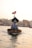 A traditional wooden boat navigates through calm waters, carrying a few passengers seated under a canopy. A flag is visible atop the boat, and the background showcases a cityscape with buildings lining the waterfront under a clear sky.