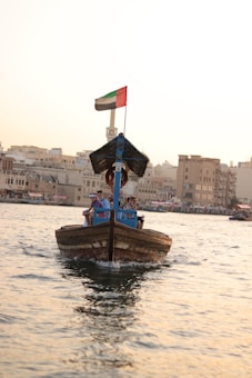 A traditional wooden boat navigates through calm waters, carrying a few passengers seated under a canopy. A flag is visible atop the boat, and the background showcases a cityscape with buildings lining the waterfront under a clear sky.