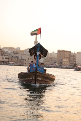 A traditional wooden boat navigates through calm waters, carrying a few passengers seated under a canopy. A flag is visible atop the boat, and the background showcases a cityscape with buildings lining the waterfront under a clear sky.