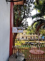 A sign indicating 'Aadhaar ATM' is mounted on a wall next to a garden area. The garden contains trees and several small potted plants organized neatly. In the background, a building with multiple windows is partially visible, and there is a row of parked motorbikes next to a fence. The scene is sunlit, giving the area a warm and welcoming feel.