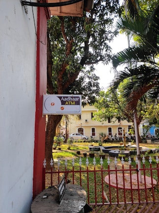 A sign indicating 'Aadhaar ATM' is mounted on a wall next to a garden area. The garden contains trees and several small potted plants organized neatly. In the background, a building with multiple windows is partially visible, and there is a row of parked motorbikes next to a fence. The scene is sunlit, giving the area a warm and welcoming feel.