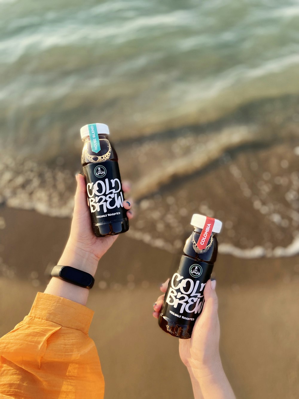 two people holding up two bottles of soda on a beach