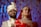 A couple dressed in traditional Indian wedding attire stands together. The woman is wearing a red outfit with intricate gold embroidery and heavy jewelry, including a nose ring and headpiece. The man is in an ornate cream-colored sherwani with a matching turban. The background features soft, blurred blue and red decorations, adding to the festive atmosphere.