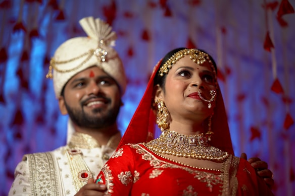 A couple dressed in traditional Indian wedding attire stands together. The woman is wearing a red outfit with intricate gold embroidery and heavy jewelry, including a nose ring and headpiece. The man is in an ornate cream-colored sherwani with a matching turban. The background features soft, blurred blue and red decorations, adding to the festive atmosphere.