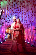 A couple dressed in traditional Indian wedding attire stands under an elaborate canopy decorated with red flowers and colorful lights. The bride wears a vibrant red lehenga with intricate embroidery, adorned with jewelry and a veil. The groom is in a white sherwani and turban, standing slightly behind her. The corridor is illuminated with multicolored lights, creating a celebratory atmosphere.