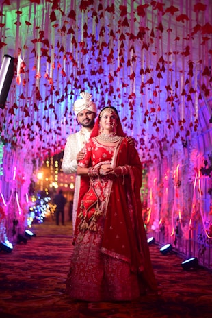 An Indian wedding couple dressed in vibrant traditional attire, smiling joyfully under a floral arch at an outdoor venue.