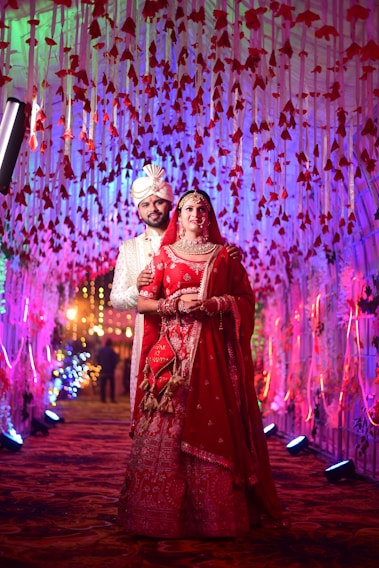A couple dressed in traditional Indian wedding attire stands under an elaborate canopy decorated with red flowers and colorful lights. The bride wears a vibrant red lehenga with intricate embroidery, adorned with jewelry and a veil. The groom is in a white sherwani and turban, standing slightly behind her. The corridor is illuminated with multicolored lights, creating a celebratory atmosphere.
