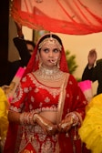 A bride dressed in a traditional red and gold lehenga, adorned with intricate jewelry including a maang tikka, necklace, earrings, and nose ring. Her hands are decorated with henna designs, and she stands under a red veil held by attendants. The background contains blurred figures and vibrant colors.