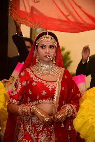 A bride dressed in a traditional red and gold lehenga, adorned with intricate jewelry including a maang tikka, necklace, earrings, and nose ring. Her hands are decorated with henna designs, and she stands under a red veil held by attendants. The background contains blurred figures and vibrant colors.