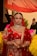 A bride dressed in a traditional red and gold lehenga, adorned with intricate jewelry including a maang tikka, necklace, earrings, and nose ring. Her hands are decorated with henna designs, and she stands under a red veil held by attendants. The background contains blurred figures and vibrant colors.