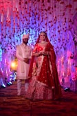 A couple dressed in traditional Indian wedding attire stands under a decorated canopy with red and white flowers and hanging ribbons. The bride wears a red lehenga and intricate jewelry, while the groom wears a white sherwani and turban. Colorful lights and floral arrangements create a festive atmosphere.