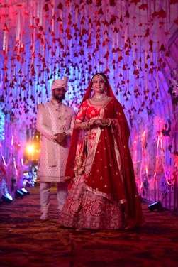 A couple dressed in traditional Indian wedding attire stands under a decorated canopy with red and white flowers and hanging ribbons. The bride wears a red lehenga and intricate jewelry, while the groom wears a white sherwani and turban. Colorful lights and floral arrangements create a festive atmosphere.
