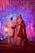 A couple dressed in traditional Indian wedding attire stands under a decorated canopy with red and white flowers and hanging ribbons. The bride wears a red lehenga and intricate jewelry, while the groom wears a white sherwani and turban. Colorful lights and floral arrangements create a festive atmosphere.