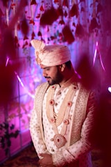 A man dressed in ornate traditional attire stands with his hands clasped. The outfit features intricate embroidery, pearl-studded jewelry, and a matching elaborate turban. The background is filled with hanging flower decorations, illuminated with purple and pink lighting, creating a festive and celebratory atmosphere.