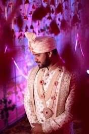 A man dressed in ornate traditional attire stands with his hands clasped. The outfit features intricate embroidery, pearl-studded jewelry, and a matching elaborate turban. The background is filled with hanging flower decorations, illuminated with purple and pink lighting, creating a festive and celebratory atmosphere.