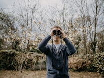 A person stands outdoors in a natural setting, holding binoculars up to their face, surrounded by flowering trees and bushes. The scene suggests early spring with budded branches and blooming flowers.